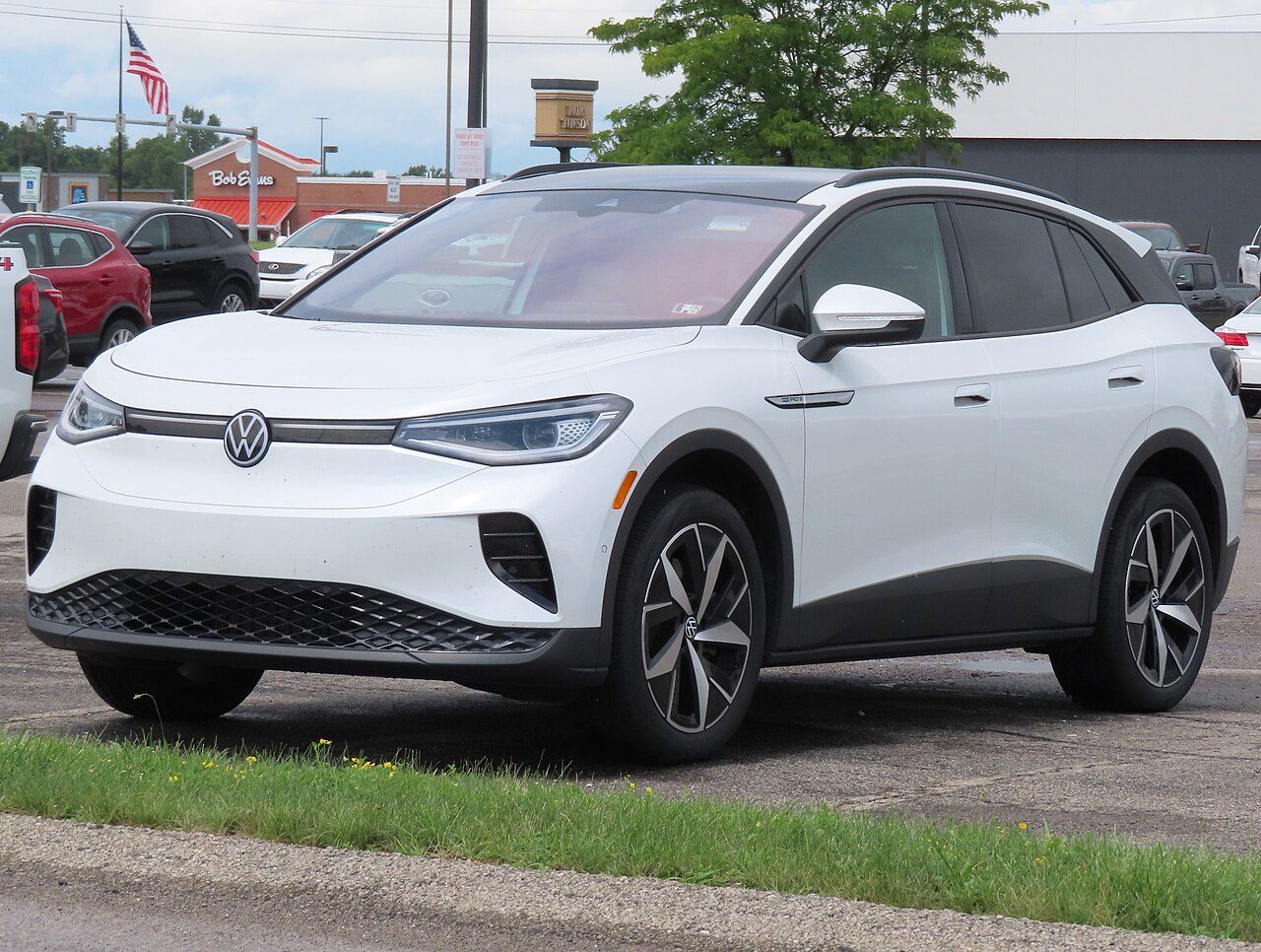 A white Volkswagen ID.4 electric SUV with a black roof and two-tone wheels, parked on asphalt beside a grass strip, with other cars and a restaurant behind it