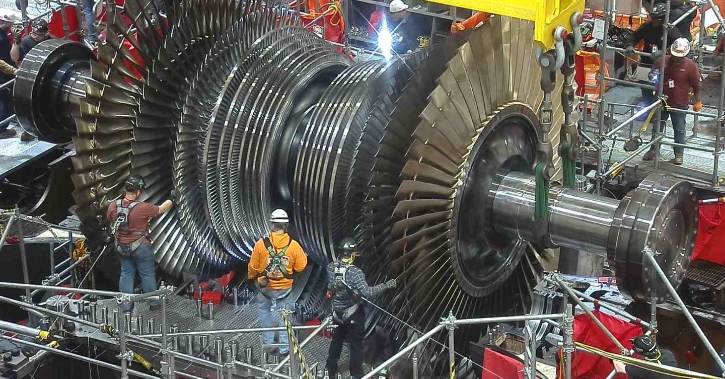 Workers in hard hats and harnesses stand on scaffolding around an opened steam turbine rotor with rows of curved blades, with a welding spark visible behind it at the top of the frame