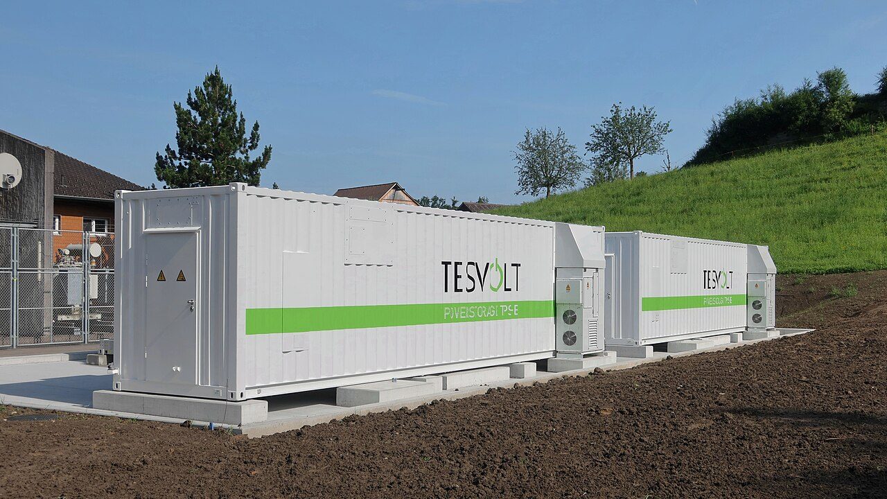 Two white Tesvolt shipping-container battery units with green stripes on concrete footings beside bare soil, a grassy hillside and trees behind them