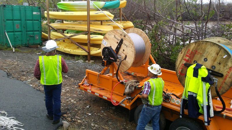 Two linemen in high-visibility vests work at an orange trailer carrying two wooden reels of conductor, with yellow kayaks stacked on a rack behind them