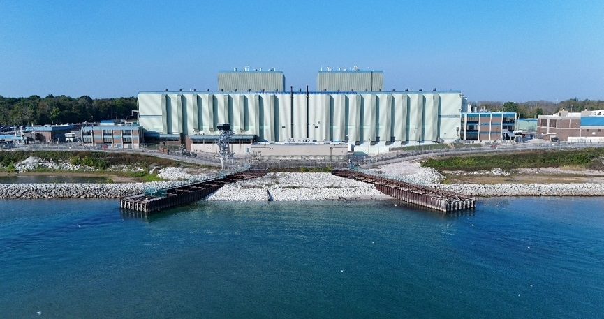 Aerial view from the water of the Point Beach Nuclear Plant: a long pale green building with two rooftop structures, two dark piers extending into blue water, white rock along the shore, lower buildings at each end and trees behind under a clear sky