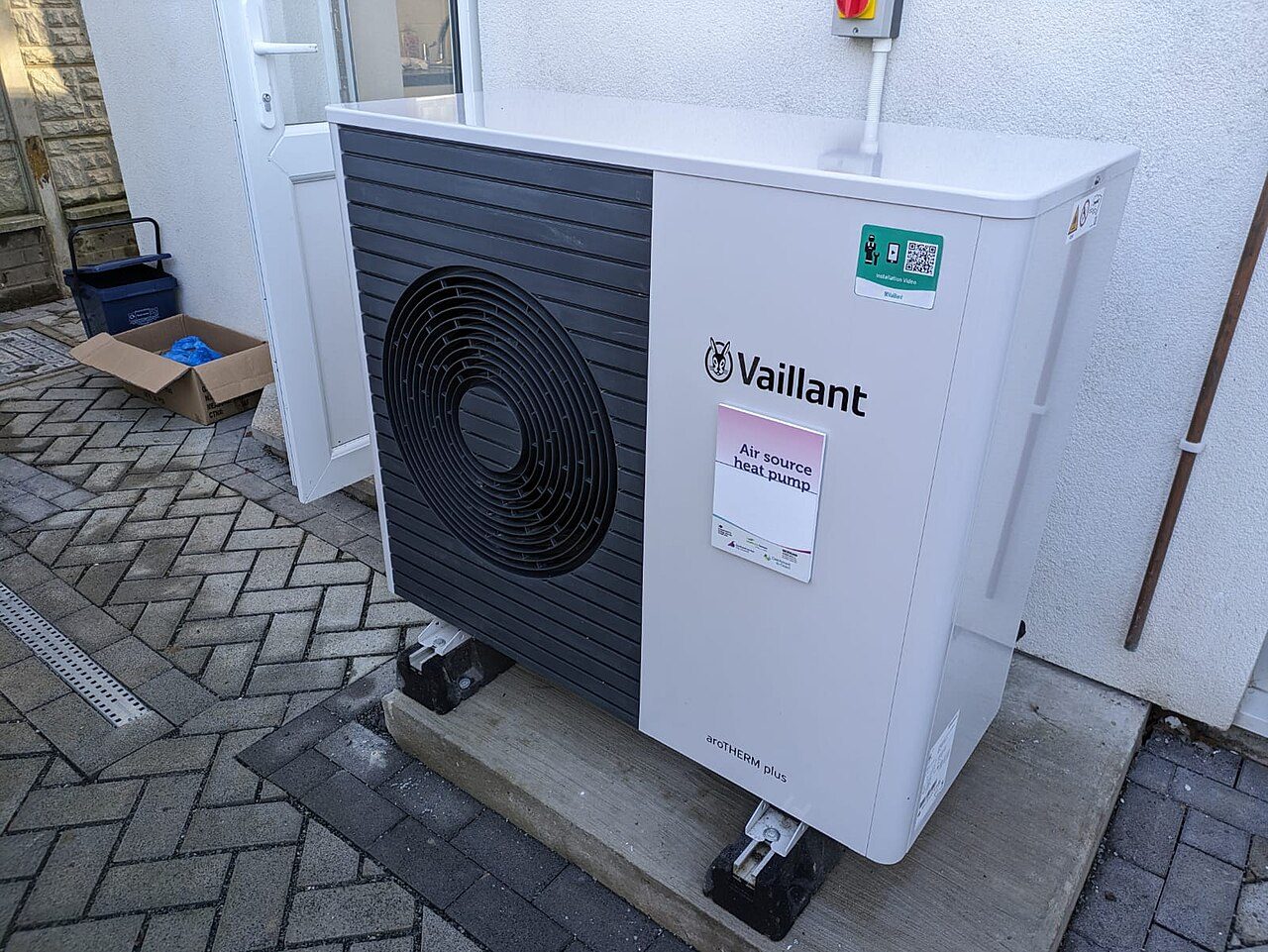 A white Vaillant aroTHERM plus air source heat pump with a dark grey fan grille stands on a concrete plinth on a block-paved yard beside an open white back door, with a red isolator switch on the wall above it