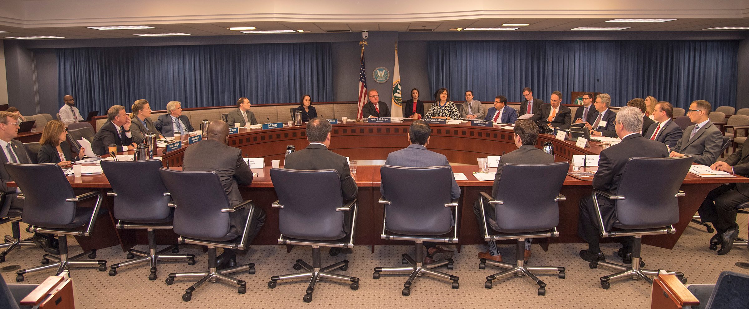 Wide view of the FERC hearing room in Washington, D.C., with about two dozen people in suits seated around a curved wooden dais with nameplates, blue curtains, and the US flag and FERC seal on the wall behind the chairman's seat