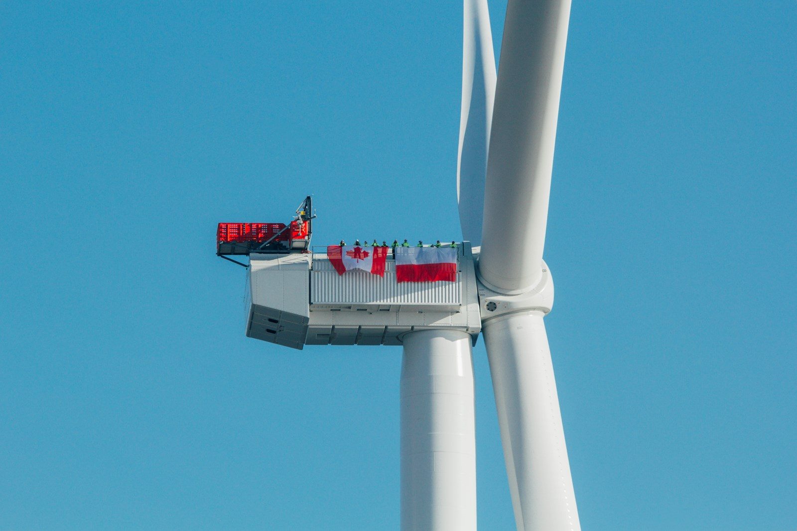 A Baltic Power wind turbine nacelle with Canadian and Polish flags hung along its side and a row of workers standing on top, against a clear blue sky